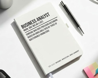 May include: A white notebook with the text "BUSINESS ANALYST" and a humorous definition. The image also includes a cup of coffee, glasses, pens, and sticky notes, suggesting an office or workspace setting.