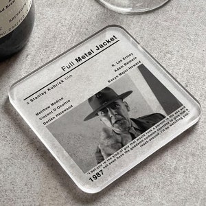 May include: A clear glass coaster with a black and white photo of a man in a military uniform. The text "Full Metal Jacket" is printed in black at the top of the coaster. The text "a Stanley Kubrick film" is printed below the title. The text "1987" is printed at the bottom of the coaster.