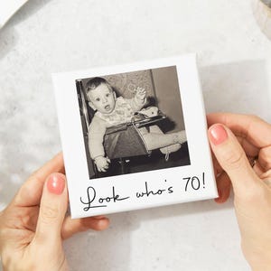Pode incluir: Impress&atilde;o em tela quadrada branca com uma foto a preto e branco de um beb&eacute; numa cadeira alta. O texto "Look who's 70!" est&aacute; escrito em it&aacute;lico por baixo. Um presente sentimental.