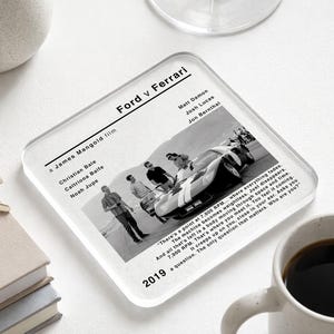 May include: A square glass coaster featuring a black and white print of the film Ford v Ferrari. The design includes the movie title, cast names, director credit, a scene with a race car and people, the year 2019, and a quote about 7,000 RPM.