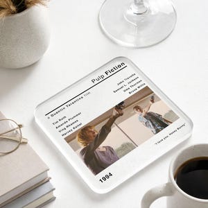 May include: A square glass coaster featuring a movie poster design for "Pulp Fiction." The coaster includes the film's title, cast, and the year 1994. The image shows a scene from the movie with two characters holding guns.