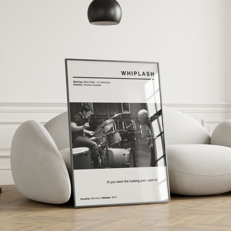 May include: A framed black and white movie poster for the film "Whiplash" leans against a white chair. The poster features the film's title, credits, and a still image of a drummer. The text "/if you want the fucking part, earn it!" is also visible.
