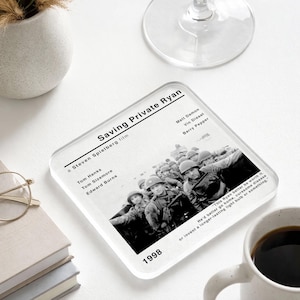 May include: Square coaster featuring a black and white image from the movie "Saving Private Ryan." The coaster includes the film's title, cast, and the year 1998. A quote is printed at the bottom. The coaster is on a white surface.