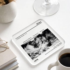May include: Square glass coaster featuring a black and white image from the 1979 film "Alien." The coaster includes the film title, cast names, and a quote. The coaster is on a white surface.