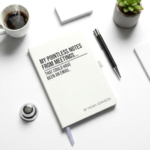 May include: A white notebook with the text "MY POINTLESS NOTES FROM MEETINGS... THAT COULD HAVE BEEN AN EMAIL." A black pen, a cup of coffee, a small succulent plant, and a silver desk bell are also on the white surface.
