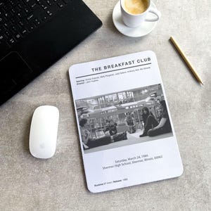 May include: A black and white mousepad featuring the movie poster for "The Breakfast Club." The mousepad includes the movie title, cast, director, and release information. A laptop, mouse, coffee cup, and pencil are also visible.