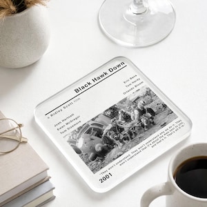 May include: Square glass coaster featuring the movie poster for "Black Hawk Down." The coaster has a black and white image of a helicopter and soldiers. The movie title, cast, and a quote are printed on the coaster.