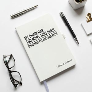 May include: A white notebook with the text "MY BRAIN HAS TOO MANY TABS OPEN SOMEBODY PLEASE SEND HELP!" and the name "NOAH JOHNSON" printed on the cover. A silver pen, a black pen, and black glasses are also visible.