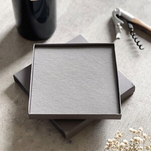 Peut inclure: Une bo&icirc;te cadeau carr&eacute;e grise avec un couvercle assorti, pos&eacute;e sur une bo&icirc;te similaire. Une bouteille de vin et un tire-bouchon sont visibles en arri&egrave;re-plan. La bo&icirc;te est probablement destin&eacute;e &agrave; de petits cadeaux ou articles de vente au d&eacute;tail.