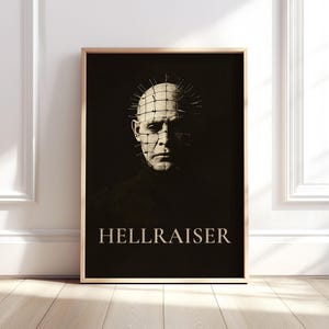 May include: Framed poster of a character from "Hellraiser", with pins in their head. The image is monochrome, with the title "HELLRAISER" in white at the bottom. The poster is displayed in a wooden frame, suitable for a horror film enthusiast.
