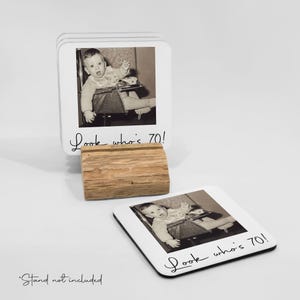 Pode incluir: T&aacute;bua de copos quadrada branca com borda preta, cada uma com uma foto vintage em preto e branco de um beb&ecirc; em uma cadeira alta. O texto "Look who's 70!" est&aacute; escrito em it&aacute;lico. Inclui um suporte de madeira.