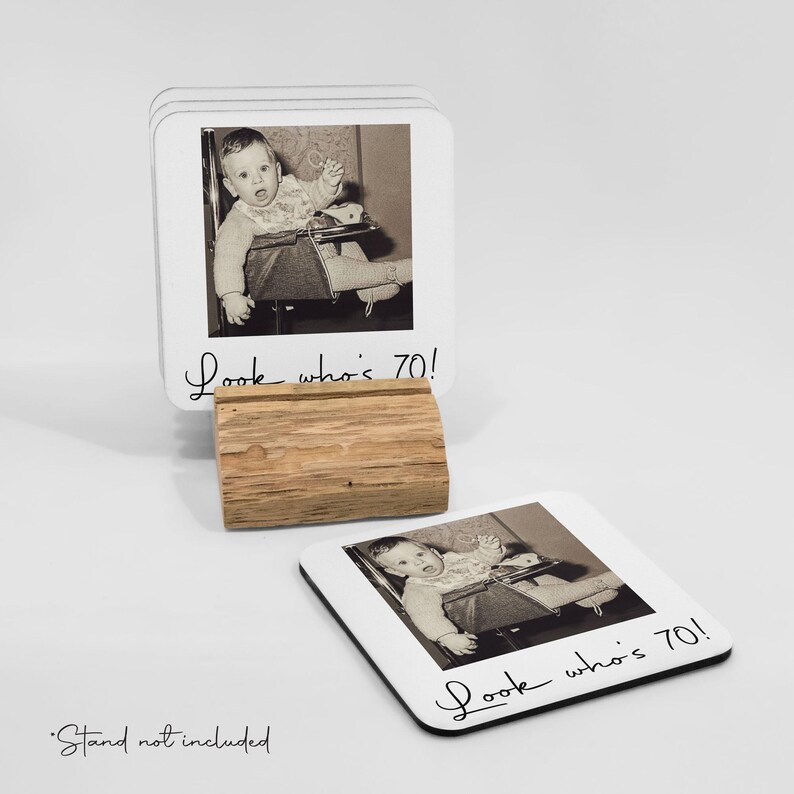 Pode incluir: T&aacute;bua de copos quadrada branca com borda preta, cada uma com uma foto vintage em preto e branco de um beb&ecirc; em uma cadeira alta. O texto "Look who's 70!" est&aacute; escrito em it&aacute;lico. Inclui um suporte de madeira.