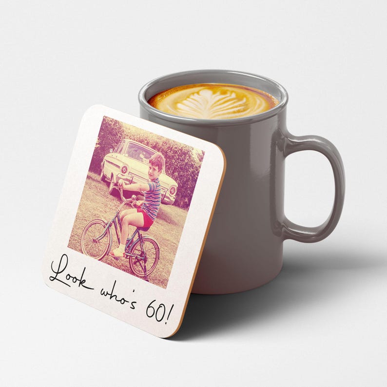 Pode incluir: Uma caneca cinza cheia de caf&eacute; e uma base para copos. A base para copos apresenta uma foto vintage de uma crian&ccedil;a de bicicleta com o texto "Look who's 60!". A foto tem uma est&eacute;tica retr&ocirc;, com um carro ao fundo.