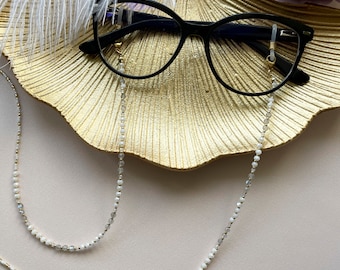 May include: A pair of black cat-eye glasses with a white and grey beaded chain. The chain is attached to the glasses with gold-toned clasps. The glasses are resting on a gold-coloured surface.