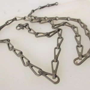 May include: A metallic chain featuring elongated, triangular-shaped links connected in a repeating pattern. The chain has a matte, dark grey finish and includes a small, circular clasp mechanism at one end for securing the links together.