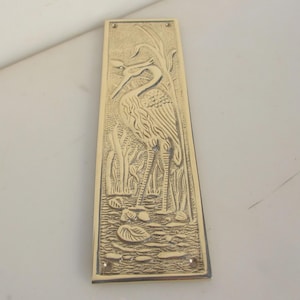 May include: A rectangular brass door plate with a detailed relief of a heron in a marsh setting. The heron is depicted amongst reeds and lily pads. The plate has a polished finish and mounting holes. The design is in a relief style.