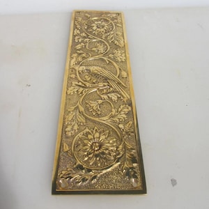 May include: A rectangular, gold-coloured metal decorative plate with an embossed design. The design features a bird perched on a branch, surrounded by floral and leaf patterns. The plate has a textured background and a polished border.