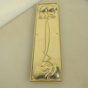 May include: A rectangular brass door plate with an Art Nouveau design. The plate features a central vertical stem with stylized floral and organic shapes at the top and bottom. The brass has a polished, reflective surface.