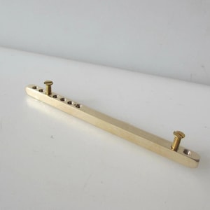 May include: A brass-coloured metal bar with multiple small holes and two screws. The bar is rectangular with rounded ends. The screws are gold-coloured and are inserted into the ends of the bar.