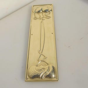 May include: A rectangular brass door plate with an Art Nouveau floral design. The plate features an embossed, stylized flower with a long stem and textured base. The brass has a polished, reflective surface.