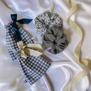 Op de afbeelding: Een zwart-witte gingham trekkoordtas met een groene strik, een zwart-witte scrunchie en een wit-zwarte scrunchie met een zwart-witte gingham print. De items liggen op een witte satijnen achtergrond.