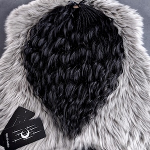 May include: Black wavy hair extensions with a braided top section, resting on a gray faux fur surface. A black and white card with a crescent moon and star design is visible in the lower left corner.