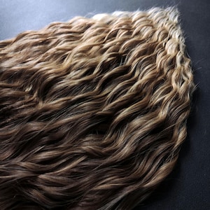 K&ouml;nnte beinhalten: Eine lange, gewellte Haarverl&auml;ngerung mit einem Ombre-Farbverlauf von hellbraun zu blond.