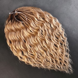 Op de afbeelding: Een bundel golvende, blonde synthetische hairextensions. Het haar is een mix van lichte en medium blonde tinten, met een donkerdere wortelzone. De extensions zijn samengebonden met een wit lint. Het haar heeft een natuurlijke, getextureerde uitstraling.