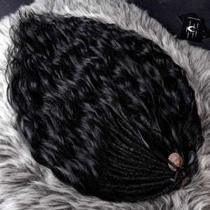May include: A bundle of black wavy hair extensions, transitioning into dreadlocks at the base. The hair is styled on a gray faux fur surface. A small tag and black ribbon are tied at the base of the dreadlocks.