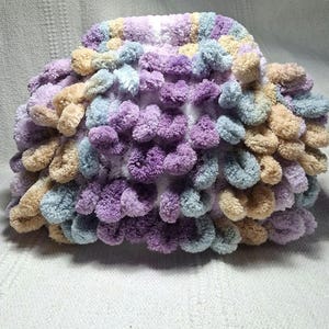 May include: A handmade, fluffy, crocheted hat in a variety of pastel colors. The hat features a textured, pom-pom-like design with shades of purple, blue, tan, and white. The soft, plush material suggests a cozy and comfortable accessory.