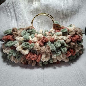 May include: A handmade, fluffy handbag in a mix of colors including cream, sage green, brown, and muted red. The bag features a gold-colored metal handle and a small leather tag. The bag is made of a soft, textured material.