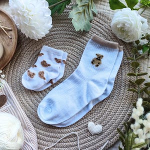 May include: A pair of baby blue socks with a brown teddy bear design, next to a smaller pair with a repeating bear pattern. The socks are on a woven mat, surrounded by white flowers and decorative elements.