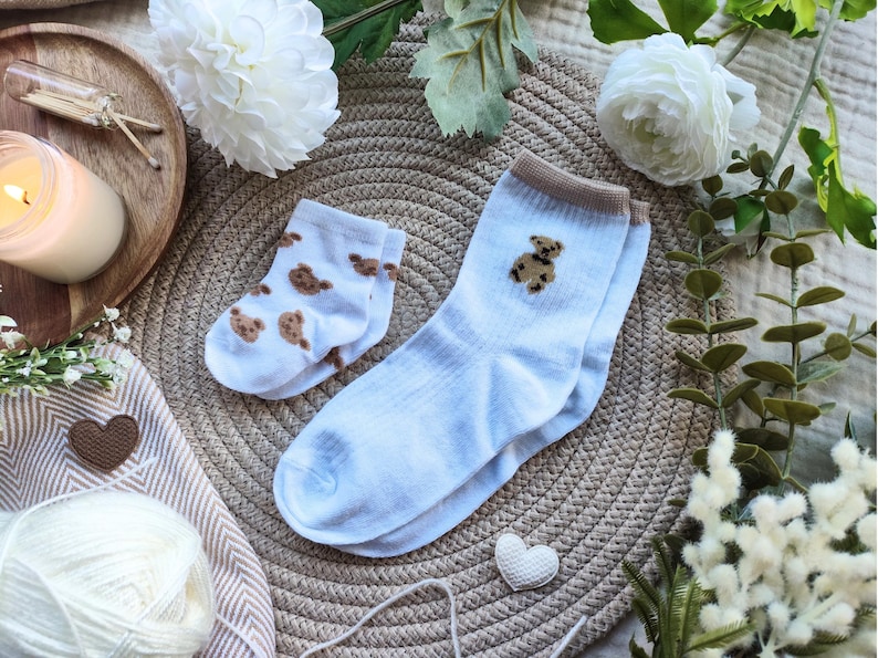 May include: A pair of baby blue socks with a brown teddy bear design, next to a smaller pair with a repeating bear pattern. The socks are on a woven mat, surrounded by white flowers and decorative elements.