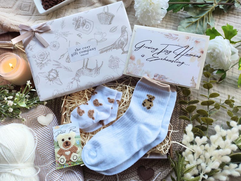 May include: A gift box with baby socks, a card, and a small teddy bear. The socks are light blue with brown teddy bear designs. The card reads "Growing Tiny Humans is Exhausting..." The gift box is wrapped in white paper with a floral design.