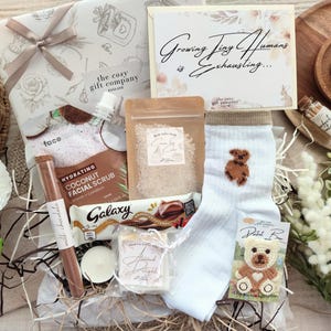 May include: A gift box with a selection of items, including a card with the text "Growing Tiny Humans is Exhausting...". The box contains a coconut facial scrub, chocolate, a candle, bath salts, a teddy bear sock, and a teddy bear patch.
