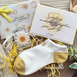 May include: A gift set featuring a daisy theme, including a gift box, card, socks, and a daisy patch. The white socks have yellow heels and toes. The gift box is wrapped with a yellow ribbon. The card reads "Something wonderful to brighten your day..."