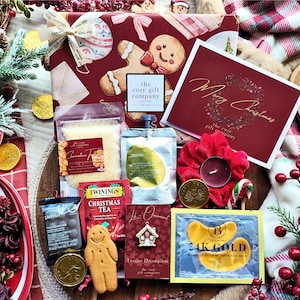 May include: A holiday gift box featuring a gingerbread man print, containing Twinings Christmas tea, a gingerbread man cookie, a 24K gold eye mask packet, a festive house ornament, a red scrunchie, a candle, and a Merry Christmas greeting card.