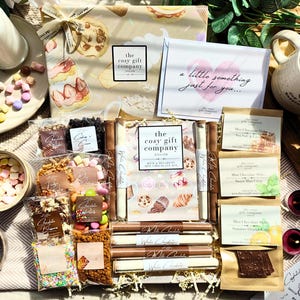 May include: A hot chocolate gift set featuring a box labelled The Cosy Gift Company, a card reading a little something just for you, various packets of chocolate toppings, chocolate stirrers, and three pouches of chocolate flakes with labels.