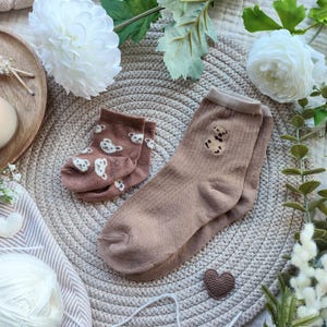 May include: Two pairs of socks in shades of brown. One pair is a light brown with white bear prints, and the other is a darker brown with a teddy bear design. The socks are displayed on a woven mat with decorative elements.