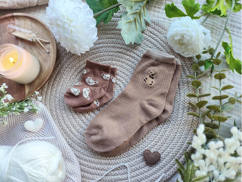 May include: Two pairs of socks in shades of brown. One pair is a light brown with white bear prints, and the other is a darker brown with a teddy bear design. The socks are displayed on a woven mat with decorative elements.