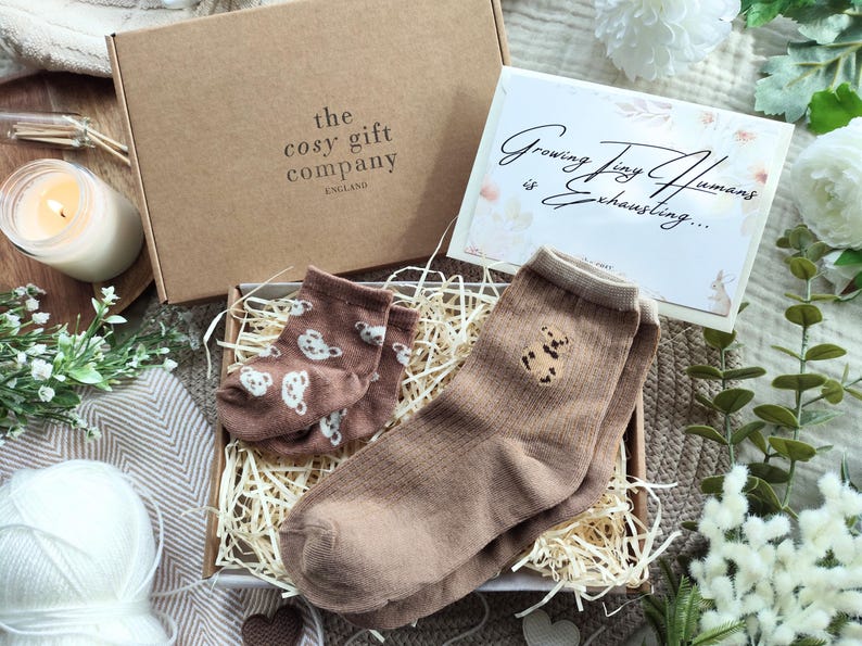 May include: A gift box from "the cosy gift company" containing a pair of brown socks with a bear design, a pair of baby socks with a bear pattern, and a card that reads "Growing Tiny Humans is Exhausting...".