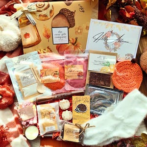 May include: A gift box from The Cozy Gift Company featuring a Hello Autumn card, various bath and food items, a crochet pumpkin, and white fuzzy socks. The box displays illustrations of cinnamon rolls, acorns, and pumpkins on a tan background.