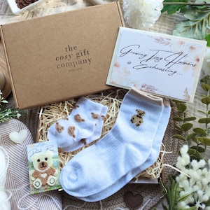 May include: A gift box from "the cosy gift company ENGLAND" contains baby socks with teddy bear designs, a card that reads "Growing Tiny Humans is Exhausting...", and a small teddy bear card. The box is surrounded by flowers and a lit candle.