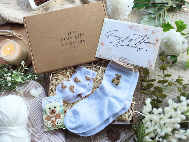 May include: A gift box from "the cosy gift company ENGLAND" contains baby socks with teddy bear designs, a card that reads "Growing Tiny Humans is Exhausting...", and a small teddy bear card. The box is surrounded by flowers and a lit candle.