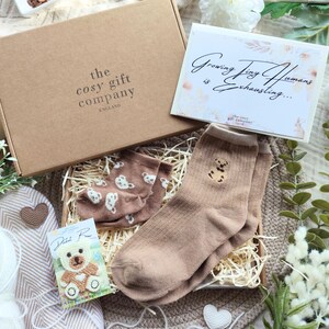 May include: A gift box from The Cosy Gift Company, England, containing a pair of tan socks with a teddy bear design, a pair of brown socks with white bear prints, and a card that reads "Growing Tiny Humans is Exhausting...".