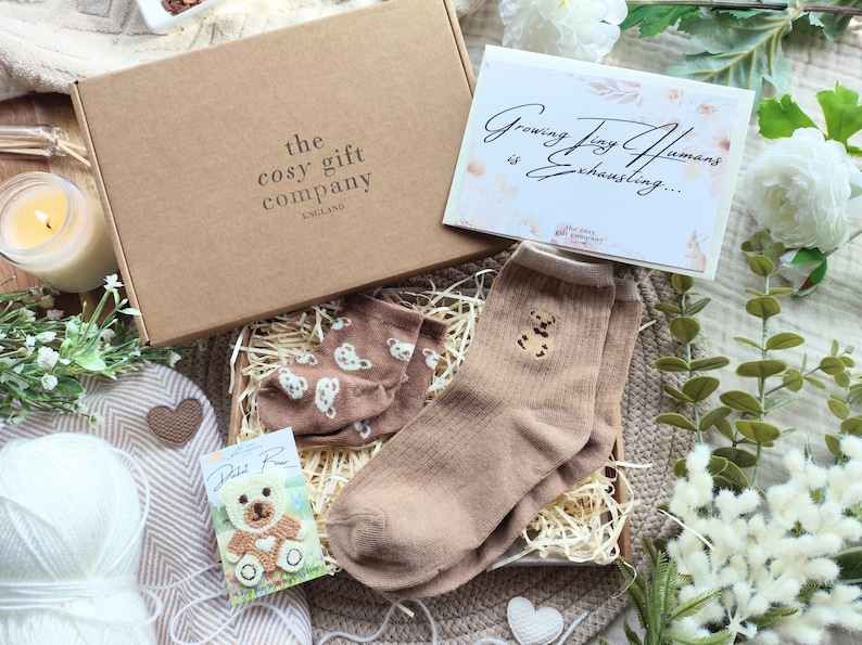 May include: A gift box from The Cosy Gift Company, England, containing a pair of tan socks with a teddy bear design, a pair of brown socks with white bear prints, and a card that reads "Growing Tiny Humans is Exhausting...".