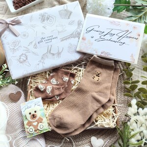 May include: A gift set with a white box tied with a ribbon, a card that reads "Growing Tiny Humans is Exhausting...", and a pair of brown socks with a teddy bear design. The set includes a small teddy bear card and a candle.
