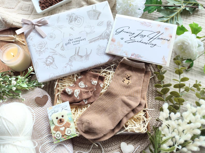 May include: A gift set with a white box tied with a ribbon, a card that reads "Growing Tiny Humans is Exhausting...", and a pair of brown socks with a teddy bear design. The set includes a small teddy bear card and a candle.