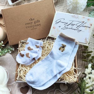 May include: A gift box from "the cosy gift company England" containing baby socks and a card that reads "Growing Tiny Humans is Exhausting...". The socks are light blue with brown teddy bear designs.