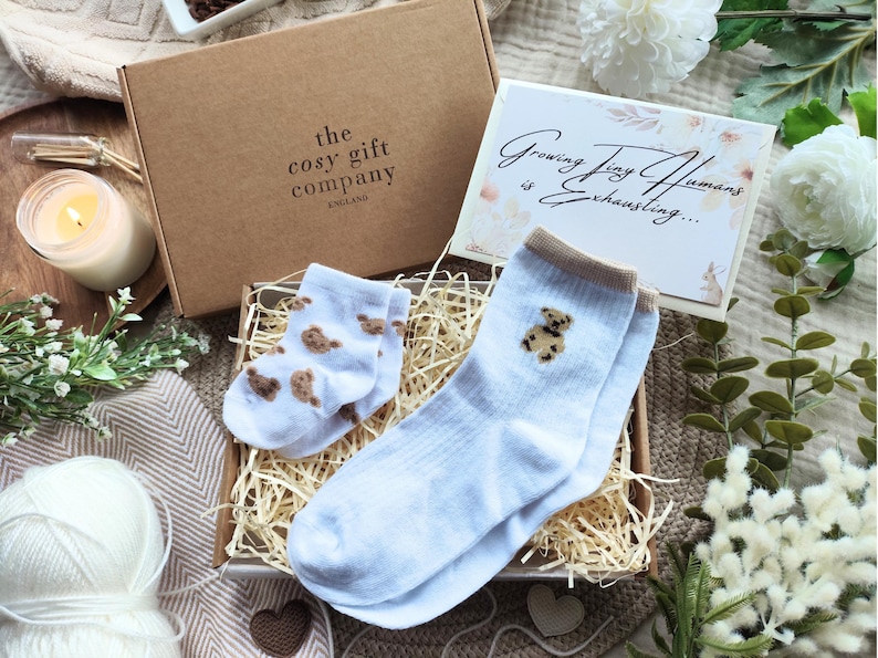 May include: A gift box from "the cosy gift company England" containing baby socks and a card that reads "Growing Tiny Humans is Exhausting...". The socks are light blue with brown teddy bear designs.